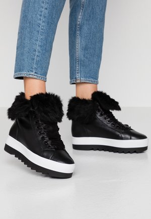 Czarne sneakersy na platformie z grubymi białymi podeszwami i czarnym futrzanym wykończeniem, noszone z krótkimi niebieskimi jeansami na jednolitym tle.
