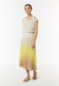 Jasno-beżowa bluzka z ukośnym wycięciem, zestawiona z plisowaną spódnicą, która przechodzi od beżu do żółci. Nosi lekkie sandały na paskach.