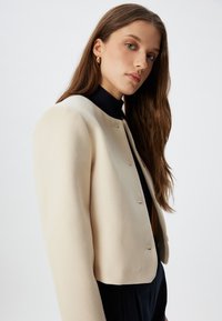 Lyhyttakki, vaalean kermanvärinen, pehmeästä kankaasta valmistettu, jossa on pyöristetyt olkapäät. Takissa on pieniä kiinnityksiä ja tummansininen kaulus alla.