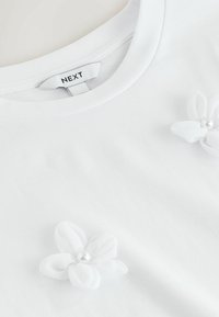 Valge ümmarguse kaelusega särk, millel on kaks valget kangasest lille, kummalgi väike keskmine pärl, ning nähtav rõivamärgis, millel on kiri "NEXT".