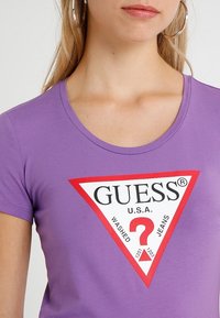 Paarse T-shirt met korte mouwen met een groot rood en wit driehoekig logo met de teksten "GUESS" en "WASHED JEANS". Zachte, getailleerde stof.