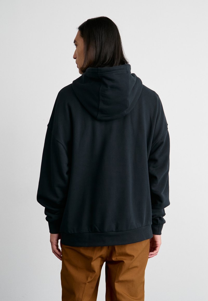 Haut à capuche noir avec une grande capuche ajustable, coupe décontractée et poignets côtelés, associé à un pantalon marron. Le tissu semble doux et lisse.
