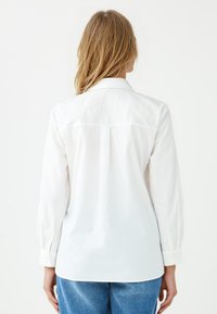 White long-sleeve shirt with a collar, back yoke seam, buttoned cuffs, and a relaxed fit. Soft, smooth fabric with a subtle sheen.