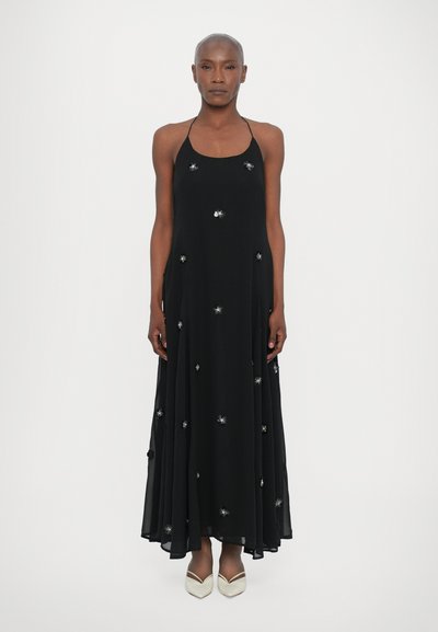 Black halter maxi dress with silver embellishments. Flowing fabric, A-line shape, and low back design. Worn with cream-colored pointed shoes.