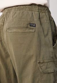 Plan rapproché d’un pantalon cargo vert olive avec une ceinture élastique, une poche passepoilée arrière, une poche latérale à rabat et une petite étiquette de marque noire.