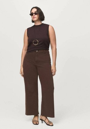 Top sin mangas marrón combinado con pantalones amplios marrones, con un dobladillo sin rematar, y sandalias negras de tiras. Acabado con un collar de colgante circular negro.