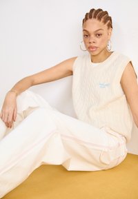 Cremfarbener, ärmelloser Pullover mit Kabelstrickmuster und blauem Logo, kombiniert mit lockeren, hellen Hosen in Off-White mit rosa Nähten und seitlichen Taschen.