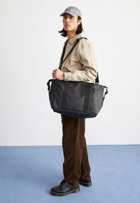 Sac de voyage en toile noire avec deux poignées épaisses et une bandoulière réglable. Caractérisé par un design élégant et minimaliste et une construction robuste.