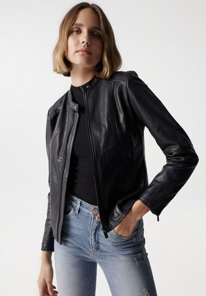 Veste en cuir noire avec fermeture éclair à l'avant, col montant, manches longues et détails de couture subtils. Portée sur un haut noir et un jean bleu.