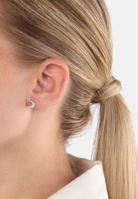 Pendiente de stud en forma de media luna plateada adornado con pequeñas piedras transparentes, con una superficie lisa y un acabado pulido.