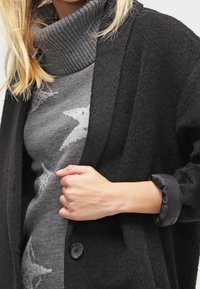 Cardigan nero in tessuto testurizzato con vestibilità ampia, dotato di colletto a scialle e due bottoni. Sotto, un maglione grigio con motivo a stelle.