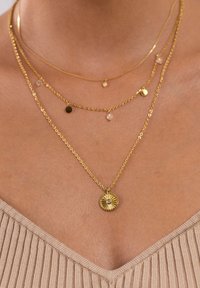 Goldfarbene, geschichtete Halsketten mit einem strukturierten, runden Anhänger, der einen zentralen Edelstein enthält. Zusätzliche zarte Ketten mit kleinen Akzenten.