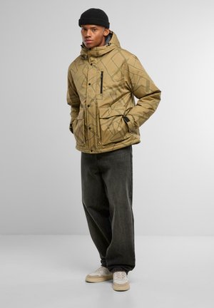 Geacă puf cu model bej, cu glugă, care dispune de un fermoar frontal și buzunare. Purtați peste blugi negri și pantofi sport crem.