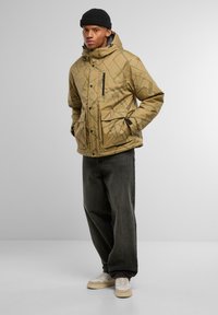 Giacca a puffo marrone con motivo, dotata di cappuccio, chiusura frontale con zip e tasche. Indossata su jeans scuri e sneakers crema.