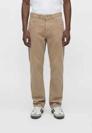 Homme portant un pantalon beige droit et des baskets blanches avec des bandes noires, debout devant un fond blanc uni.