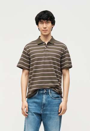 Fiatal férfi, aki olíva zöld és rózsaszín csíkos rövid ujjú galléros pólót és kék farmert visel, tiszta fehér háttér előtt áll.