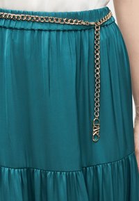 Ceinture chaîne en or avec détail de fermoir portée sur une jupe en tissu bleu sarcelle froncé à la taille.