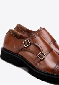 Bruine leren schoenen met twee zilveren gespen, ronde neus en zwarte rubberen zool. Gestikte details voegen textuur toe aan het gladde oppervlak.