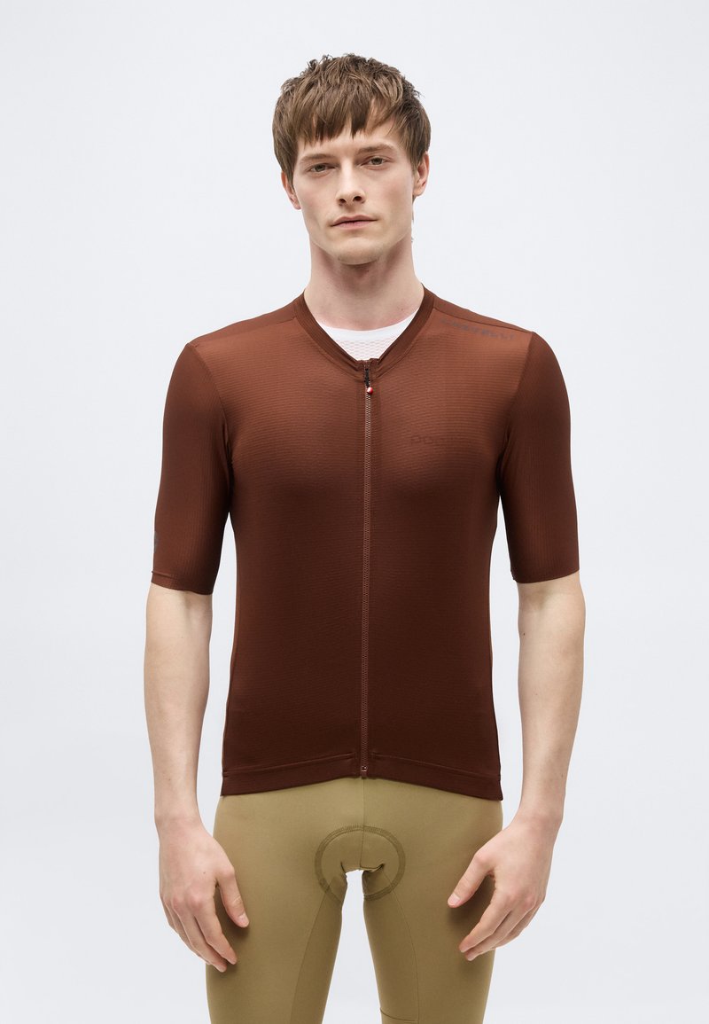 Άνδρας που φοράει στενή καφέ ζακέτα ποδηλασίας με φερμουάρ μπροστά και κοντά μανίκια, καθώς και ταμπά επενδυμένα ποδηλατικά σορτς, στέκεται μπροστά σε απλό φόντο.