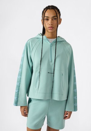 Personne aux longues tresses portant un sweat à capuche Champion bleu sarcelle clair avec des rayures logo sur les manches et un short assorti sur un fond blanc.