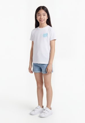 Mlada deklica stoji, oblečena v belo majico z majhnim modrim logotipom, svetlo denim kratke hlače in bele superge na ježka, rahlo se smehlja.