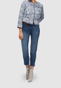 Veste en tweed bleu et blanc avec des bords effilochés et deux poches avant, associée à un jean skinny en denim foncé et des bottines gris clair.