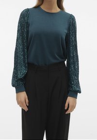 Top lungo teal con maniche a palloncino ricoperte di paillettes; tessuto morbido sul corpo; abbinato a pantaloni neri a vita alta.