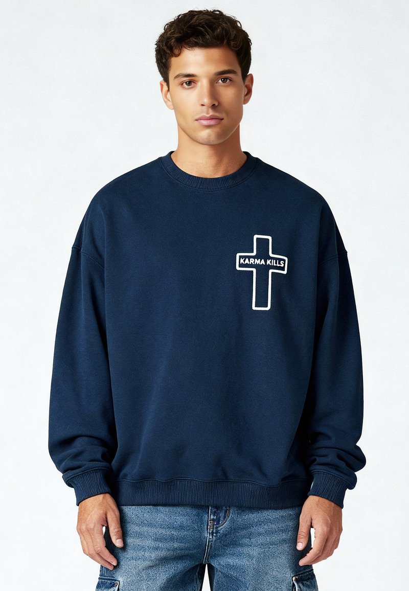 Jeune homme portant un sweat-shirt bleu marine avec une croix blanche et le texte "KARMA KILLS" sur le côté gauche de la poitrine, assorti à un jean bleu.