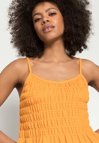 Vestido laranja com pregas, alças finas, tecido texturizado e corpete ajustado, alargando na cintura para um ajuste relaxado.