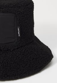 Sort bucket hat lavet af tekstureret, fuzzy materiale med en bred skygge, som har en rektangulær sort lap og et lille mærke.