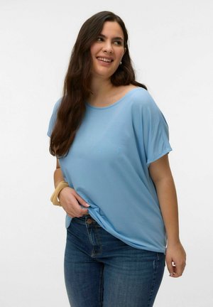 Joven con cabello largo y oscuro que lleva una blusa suelta de manga corta de color azul claro y jeans azules, sonriendo y sujetando el dobladillo de la camisa.