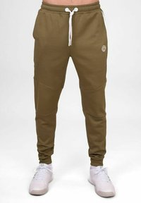 Olivgrüne Jogginghose aus Baumwollmischung mit Bindegürtel, Seitentaschen und Kniefeld-Details. Getragen mit weißen Sneakers.