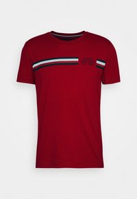 Červené tričko vyrobené z bavlny s kulatým výstřihem. Na hrudi má vodorovné pruhy v modré, bílé a tmavě zelené barvě a značkový text.