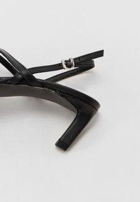 Black strappy high-heeled sandal with a square toe. Features thin crossed straps and a metallic buckle closure. Smooth leather texture.