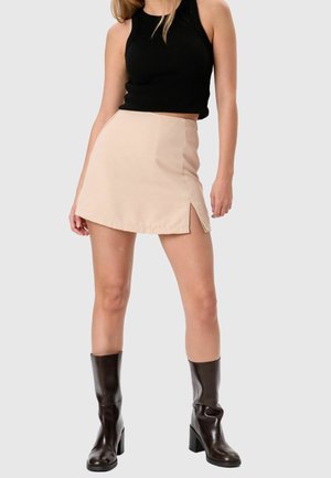 Γυναίκα φορεί μαύρο αμάνικο crop top, μπεζ μίνι φούστα με πλαϊνή σχισμή και σκούρα καφέ δερμάτινες μπότες με τακούνι μέχρι τη μέση της γάμπας, όρθια.