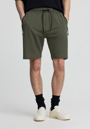 Uomo che indossa pantaloncini verde oliva con coulisse, calzini neri arricciati sopra scarpe da ginnastica bianche, in piedi con le mani in tasca.