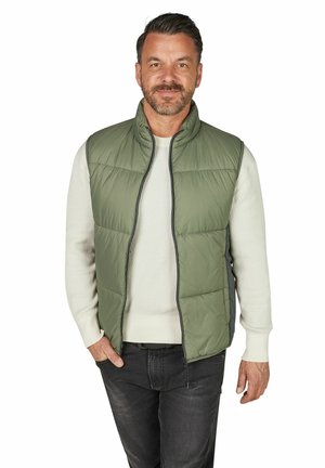 Uśmiechnięty mężczyzna ubrany w jasnozieloną pikowaną kamizelkę na białym swetrze i czarnych dżinsach, stojący na białym tle.