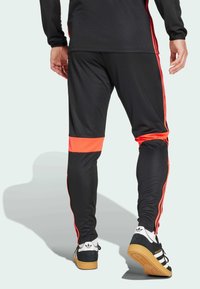 Pantalones deportivos negros con rayas laterales rojas y una banda acentuada en la parte inferior de la pierna. Combinados con zapatillas negras y blancas. Textura suave.