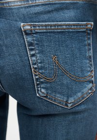 Λεπτομέρεια πίσω τσέπης μπλε τζιν με πορτοκαλί ραφές και καμπύλη διακοσμητική ραφή στην τσέπη.