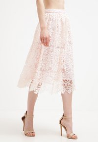 Falda midi de encaje en rosa claro, con un patrón floral, cintura elástica y dobladillo en forma de pico, combinada con sandalias de tacón nude.