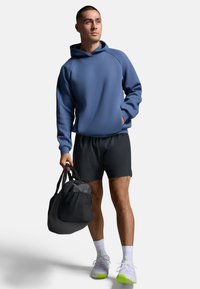 Ein Mann steht selbstbewusst in einem blauen Hoodie und schwarzen Shorts, während er eine schwarze Sporttasche hält. Er trägt weiße Turnschuhe mit grünen Akzenten.