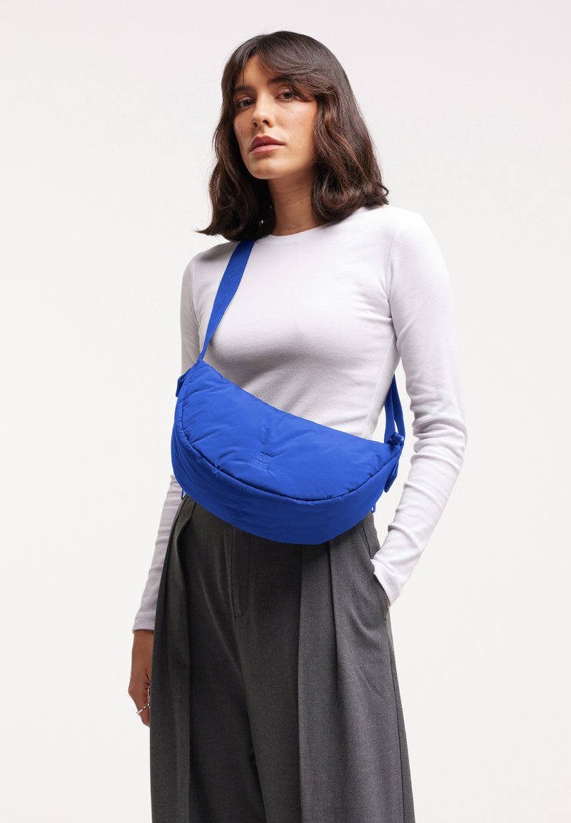 Mujer con una camiseta de manga larga blanca y pantalones grises, llevando un bolso bandolera azul brillante sobre un fondo liso.