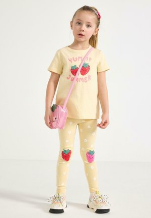 Ung pige iført en gul t-shirt og leggings med jordbærprint, der holder en lille lyserød pung formet som en jordbær, stående på et hvidt gulv.