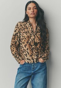 Blouse en satin beige et marron à motif léopard, avec un col noué, des boutons à l'avant et des manches longues ; associée à un jean en denim bleu.