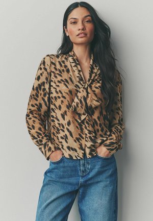 Blůza s leopardím potiskem v béžové a hnědé barvě, saténová, s uvázaným límcem, knoflíkovým zapínáním a dlouhými rukávy; kombinováno s modrými džínami.