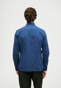 Ilgų rankovių marškiniai iš tamsiai mėlynos medžiagos, su klasikiniu apykakle, sagomis uždarytomis rankovėmis ir išlenktu apatine dalimi.