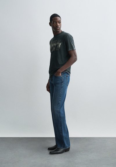 Camiseta gráfica negra con letras metálicas, combinada con jeans de denim azul de pierna recta y botas negras de punta. Diseño simple.