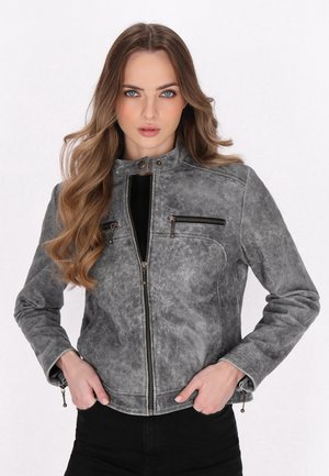 Young woman with long wavy hair wearing a gray zip-up jacket with zippered chest pockets and hands in waist pockets.