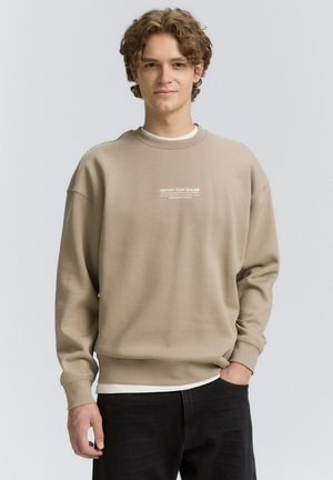 Beige sweatshirt med rund halsringning, ribbade ärmslut och en logotyp på bröstet. Bärs över en vit skjorta och kombineras med mörka jeans.
