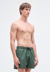 Zaļas peldbikses ar elastīgu jostu un melnu auklu. Izgatavotas no viegla materiāla, ar vienkāršu dizainu bez rakstiem.